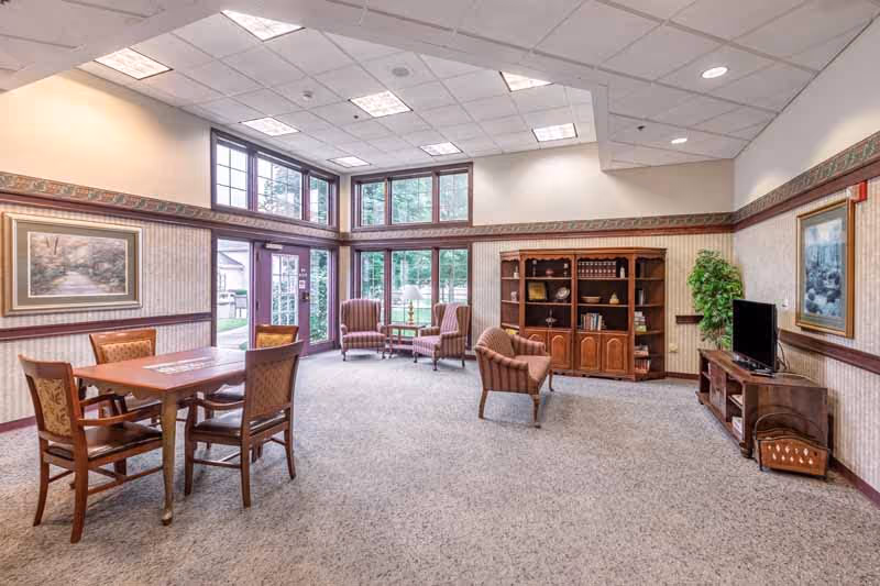 Bright communal lounge with a table and chairs, armchairs near large windows, a bookshelf and a TV.