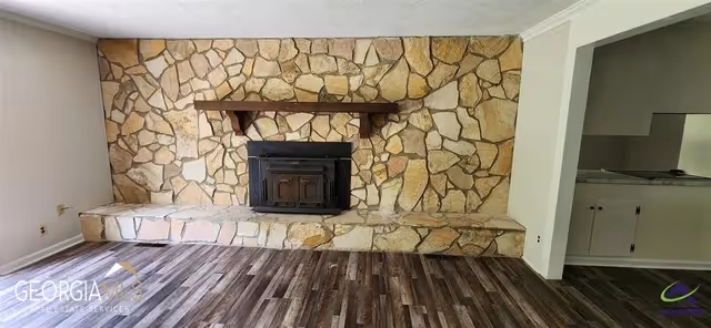 A living room with a stone fireplace centered on a stone wall. The floor is covered with dark wood laminate. To the right, there is an open doorway leading to a kitchen area with white cabinets and a countertop.