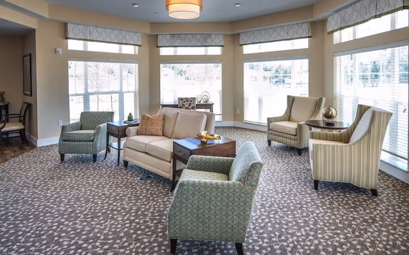 A bright and spacious living room area with large windows allowing natural light to fill the space. The room features a beige loveseat with a decorative pillow, two patterned armchairs in the foreground, and two striped armchairs near the windows. There are small wooden side tables placed between the seating, and the floor is covered with a patterned carpet. The walls are painted a soft beige color, and the windows have white blinds and valances.