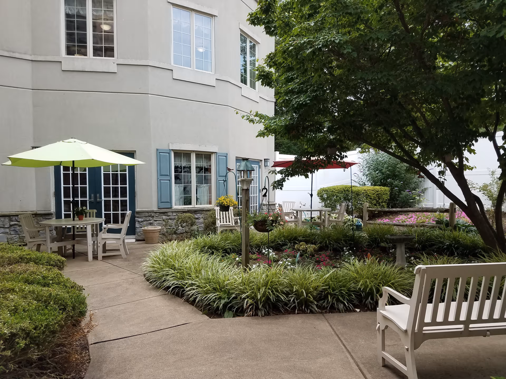 Outdoor garden area at Wyncote Place featuring a circular flower bed with various plants and flowers, surrounded by a concrete walkway. There are white benches and tables with umbrellas for shade, and the building exterior with windows and blue shutters is visible in the background.