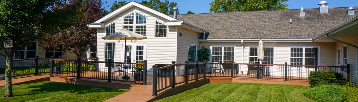 Exterior view of a single-story assisted living community building with beige siding, multiple windows, and a brown shingled roof. The building features a wooden deck with black railings, outdoor tables with umbrellas, and is surrounded by green grass and trees under a clear blue sky.