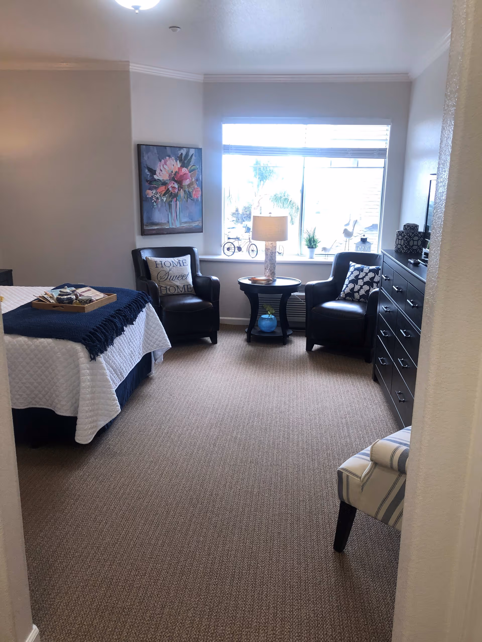 Well-lit bedroom with a bed, two armchairs flanking a window, a side table with a lamp, and a dresser.