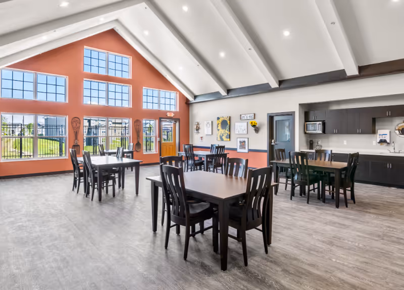 Bright communal dining room with multiple dark wooden tables and chairs, high vaulted ceiling, large windows, and a kitchenette area.