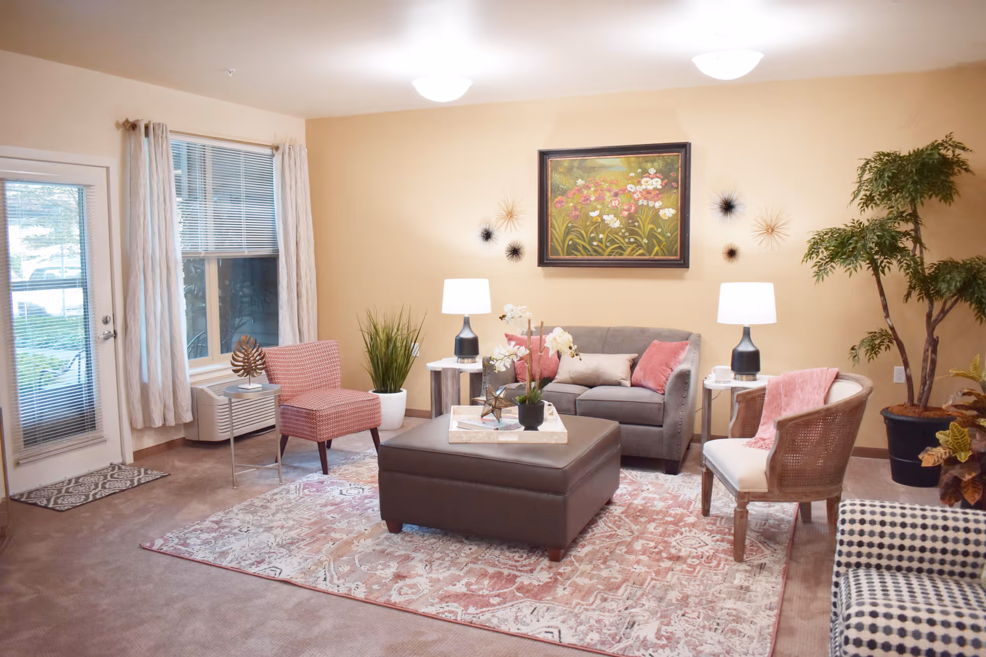 Cozy living room with a sofa, armchairs, ottoman on a patterned rug, decorative plants and wall art.