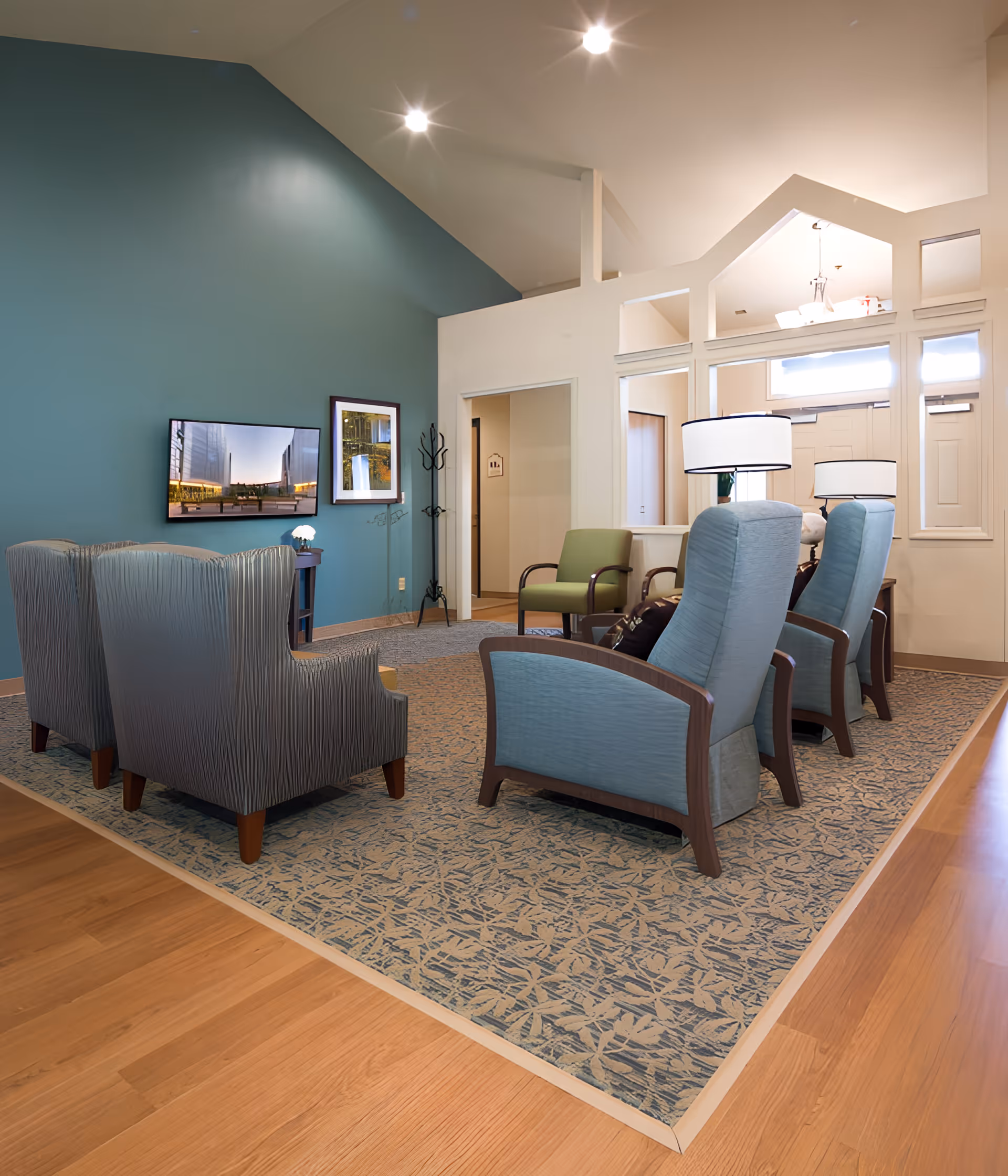 A cozy living room area in a memory care facility with a blue accent wall, a mounted flat-screen TV, framed artwork, and several upholstered chairs arranged on a patterned carpet. The room has wooden flooring, recessed ceiling lights, and a coat rack near an open doorway.