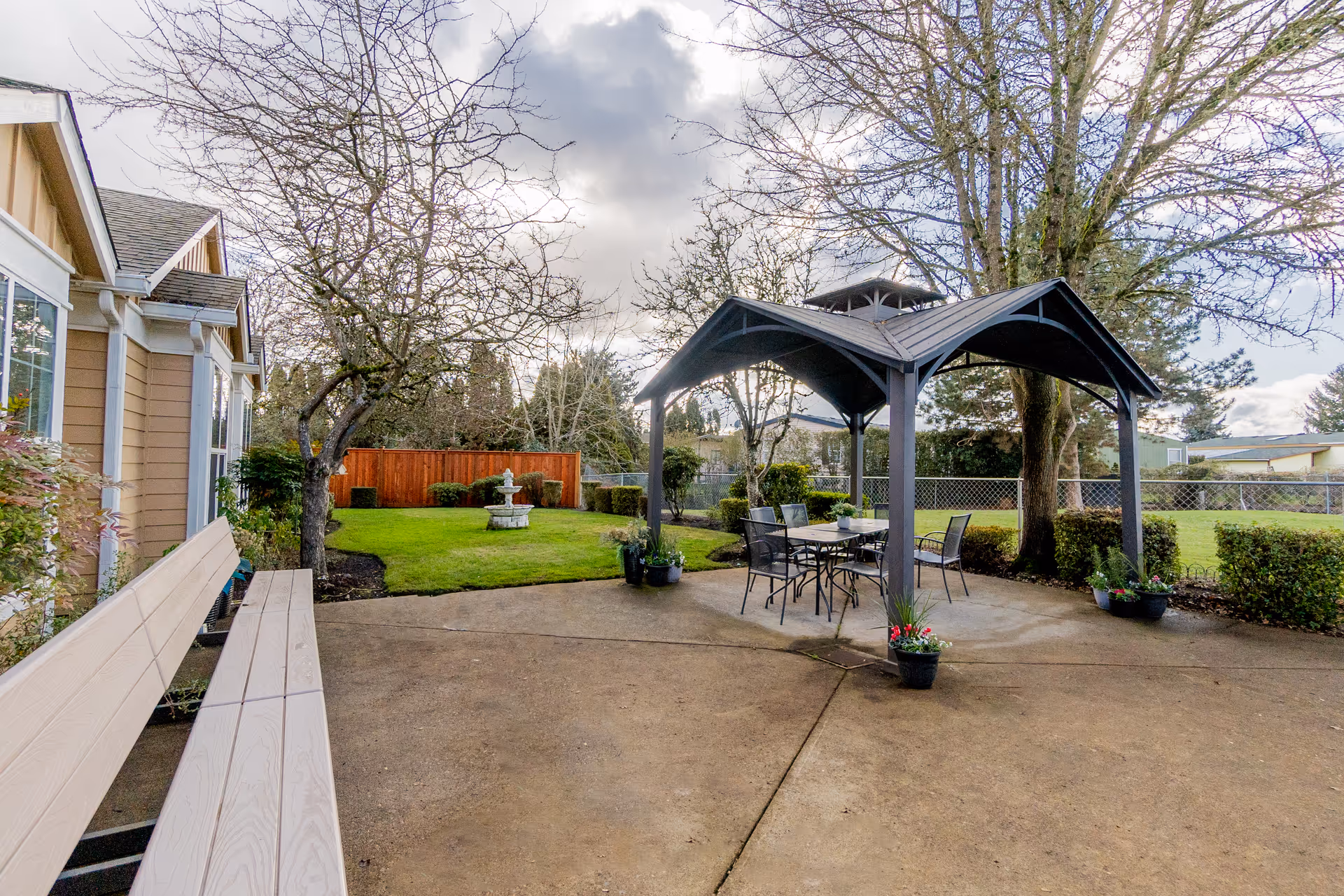 Outdoor patio with a gazebo covering a table and chairs, a nearby bench, lawn with a small fountain, and surrounding trees and fencing.