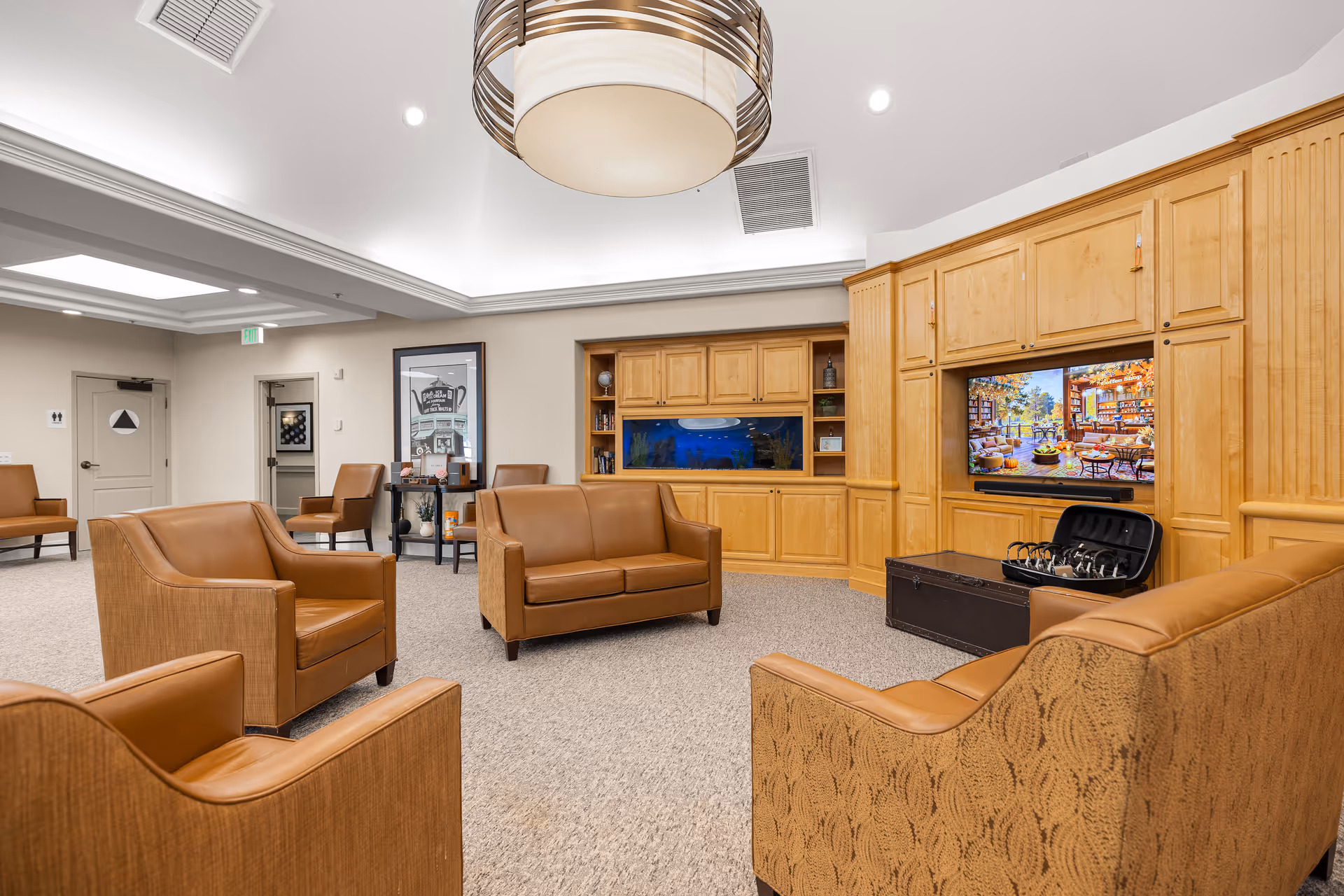 A cozy senior living facility common area with tan leather armchairs and loveseats arranged around a central space. The room features a large wooden entertainment center with a built-in aquarium and a flat-screen TV displaying a colorful indoor scene. The ceiling has recessed lighting and a modern circular pendant light fixture. The walls are light-colored, and there is a small table with decorative items and framed artwork on the wall.