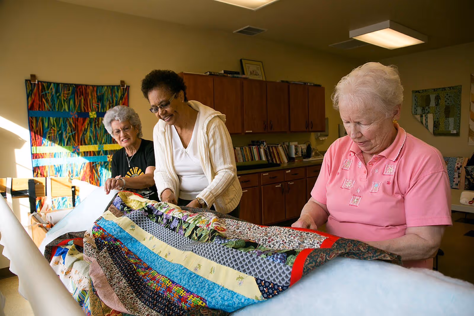 Three elderly women working together on a colorful quilt in a well-lit room with wooden cabinets and bookshelves in the background.