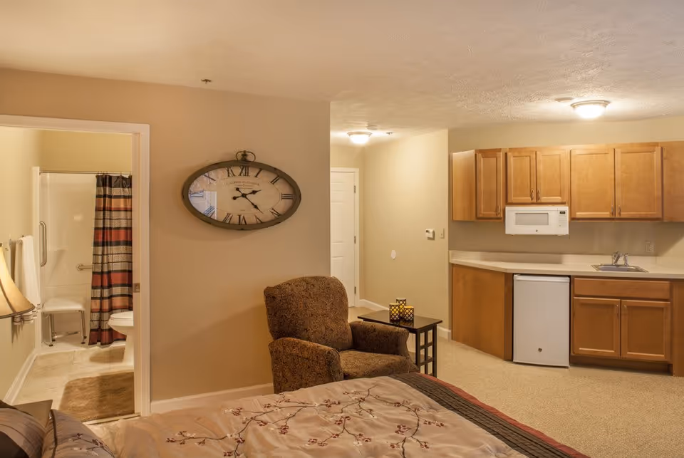 Interior view of a senior living facility room featuring a bed with floral bedding in the foreground, a patterned armchair next to a small table with decorative candle holders, a kitchenette with wooden cabinets, a microwave, a small refrigerator, and a sink. To the left, there is an open door leading to a bathroom with a shower, a shower chair, and a striped shower curtain. A large oval wall clock is mounted on the wall above the armchair.