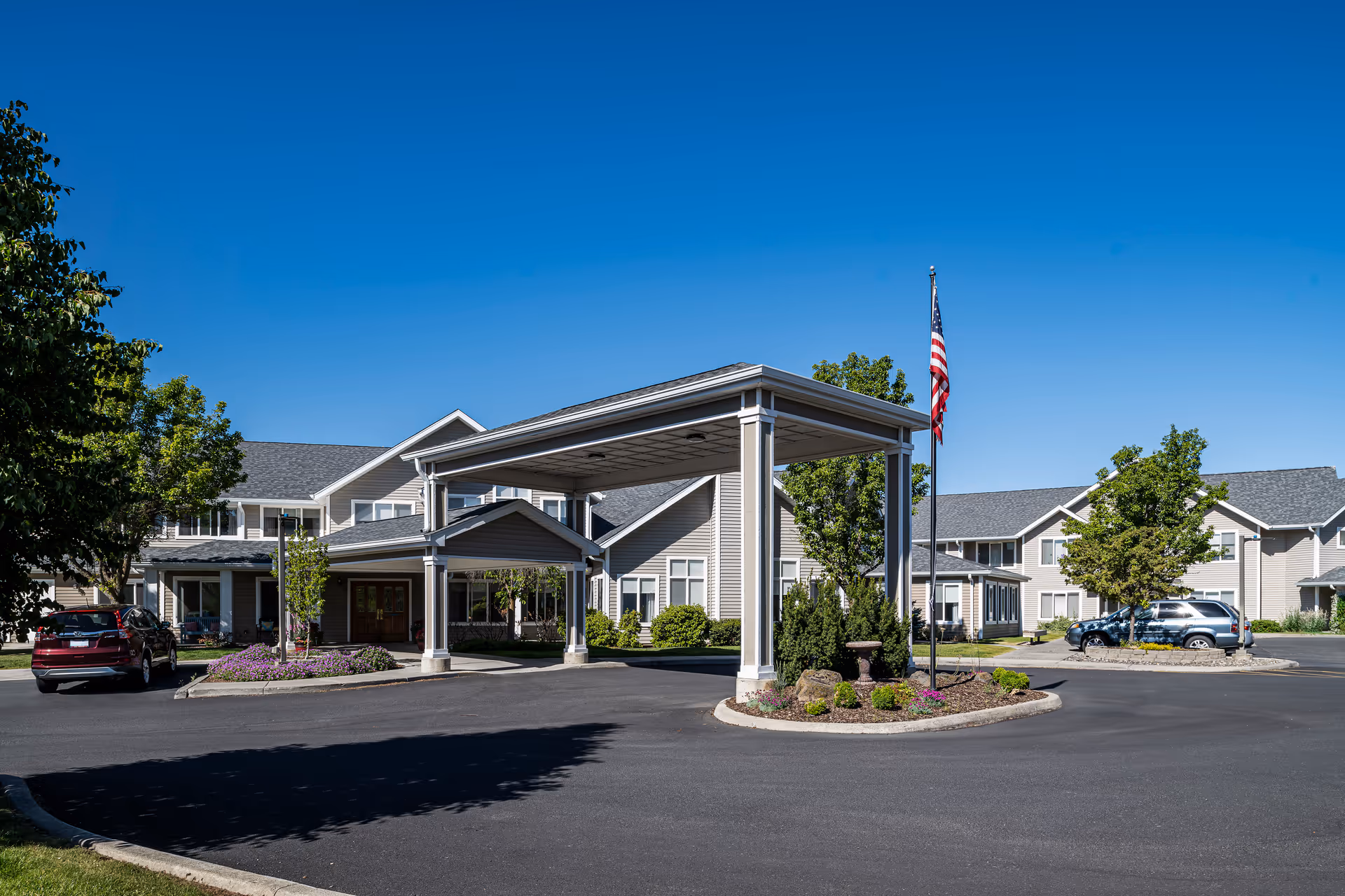 Exterior view of Bishop Place Senior Living facility showing a large covered entrance with columns, a circular driveway, landscaped areas with trees and shrubs, parked cars, and a flagpole with an American flag under a clear blue sky.