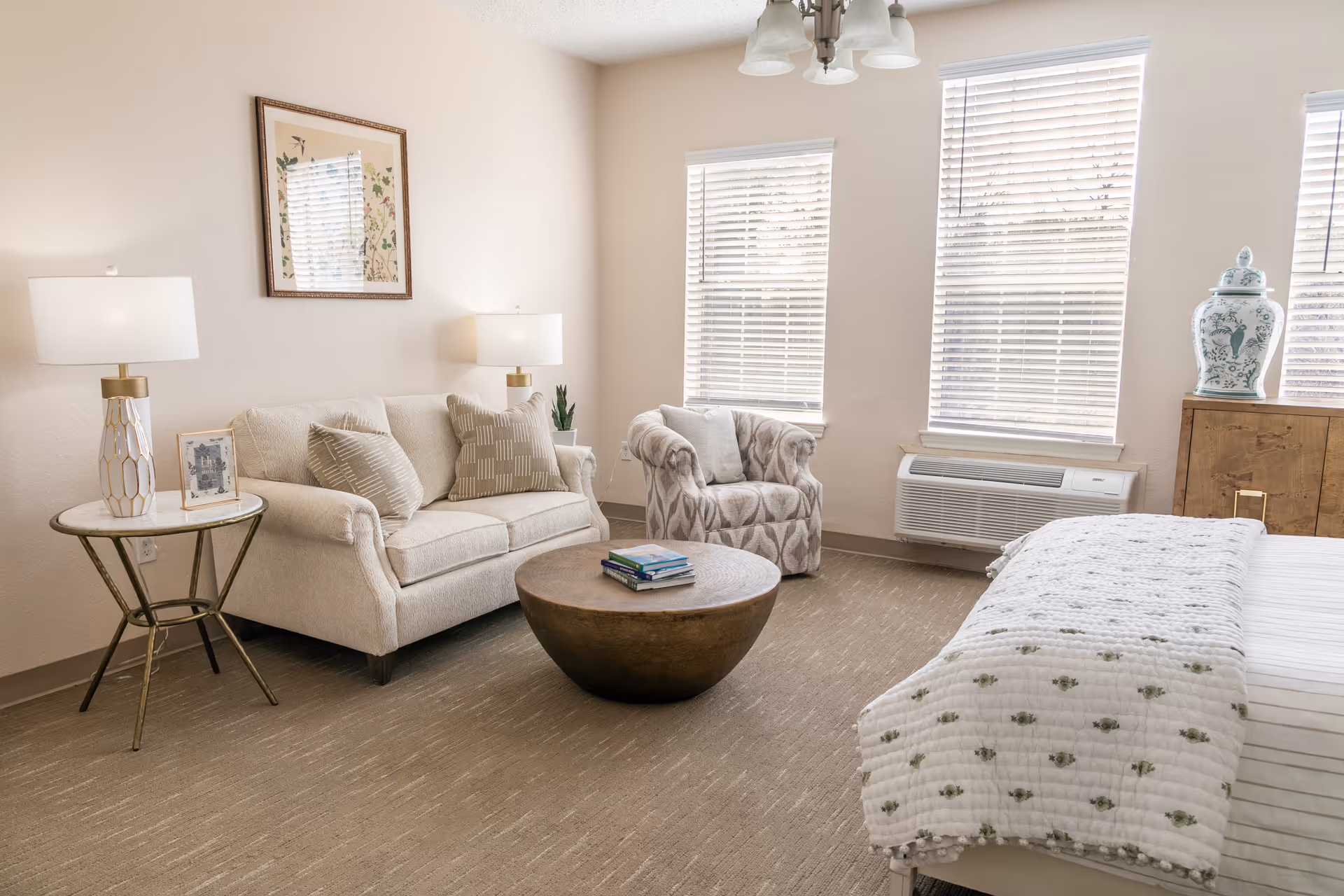 A bright and cozy senior living room featuring a beige sofa with patterned cushions, a round wooden coffee table with books on it, a patterned armchair, two table lamps on side tables, three large windows with blinds, a bed with a white and green floral quilt, and a wooden cabinet with a decorative vase.