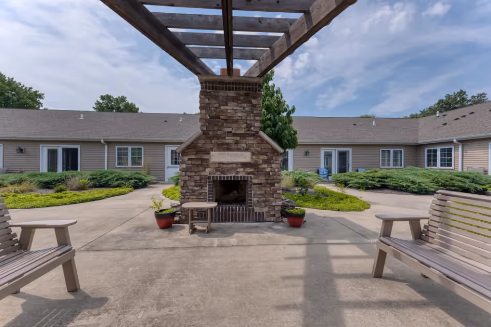 Outdoor courtyard area with a stone fireplace under a wooden pergola. There are two wooden benches facing the fireplace and potted plants on either side. The courtyard is surrounded by a single-story building with beige siding and multiple windows and doors. The sky is partly cloudy.