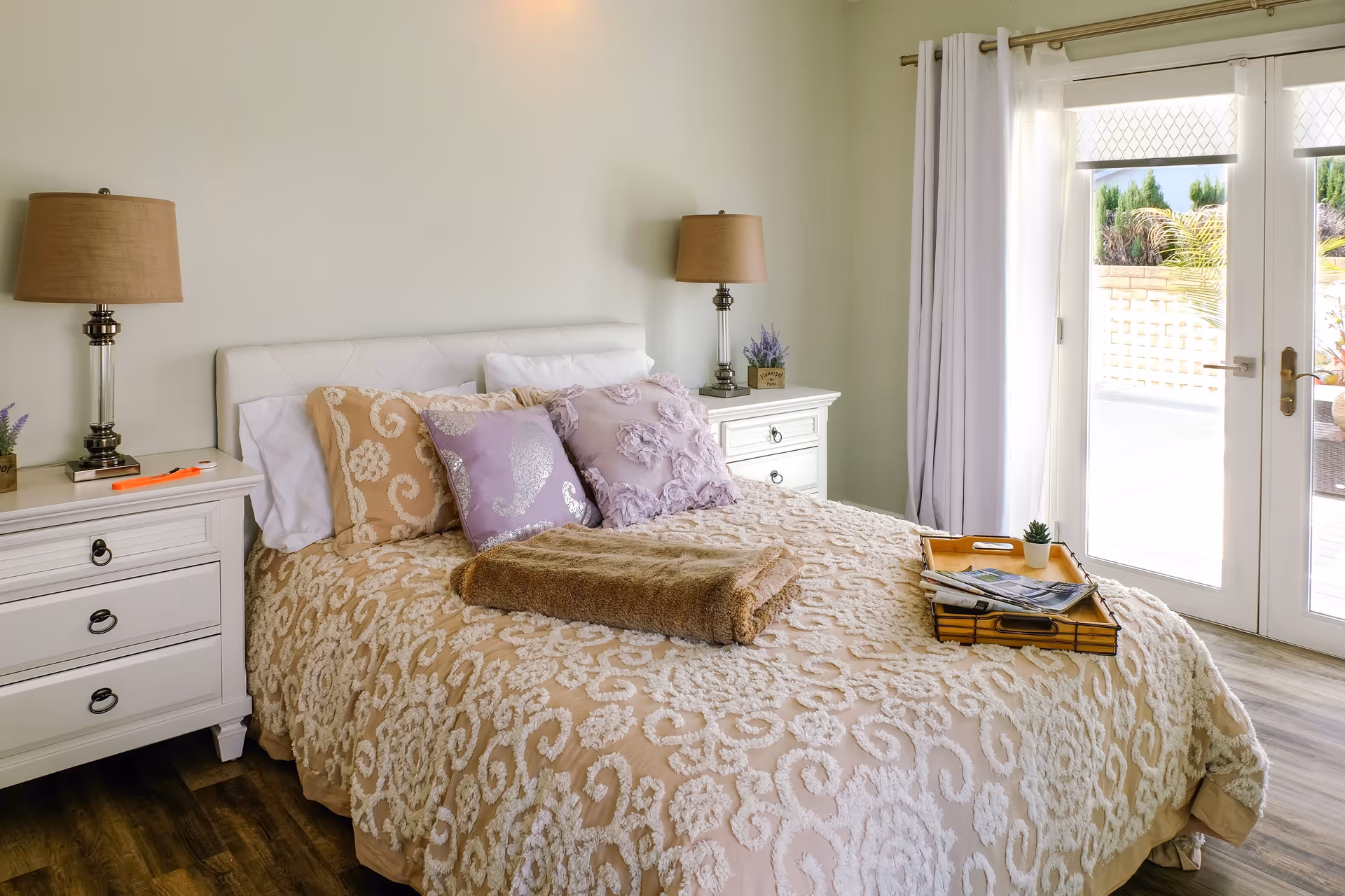 A cozy bedroom with a bed covered in a beige and white patterned bedspread, adorned with decorative pillows in beige and lavender. There are two white nightstands on either side of the bed, each with a lamp and a small potted plant. A folded brown blanket lies on the bed along with a wooden tray holding a small plant and some magazines. The room has wooden flooring and a glass door with white curtains leading to an outdoor area.