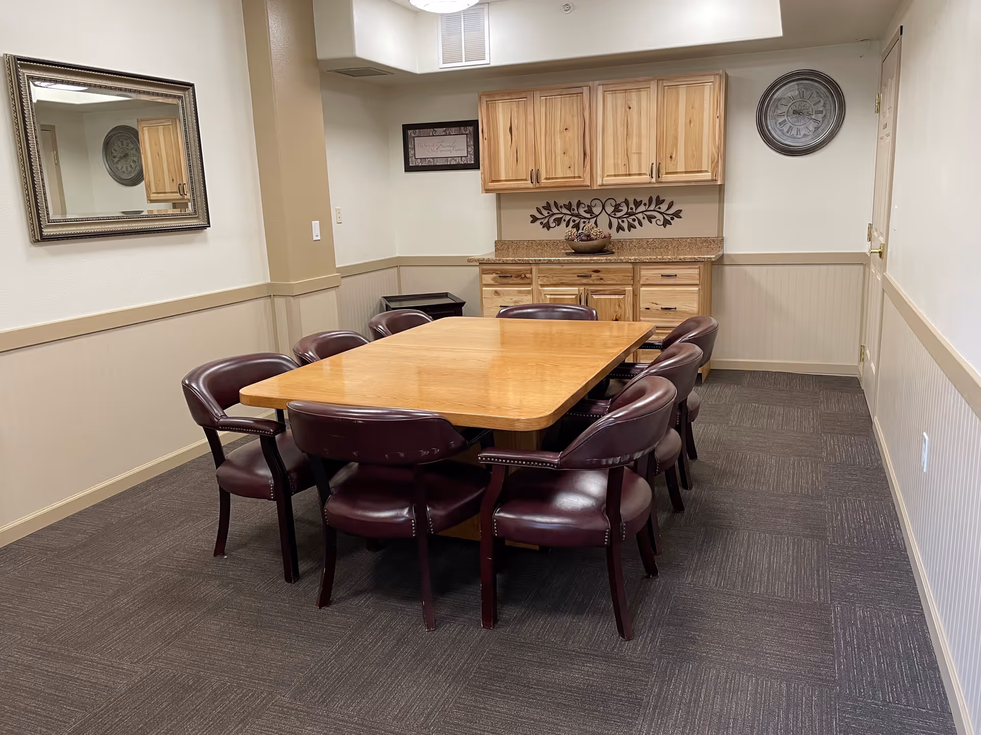 A small conference or dining room with a rectangular wooden table surrounded by eight dark brown leather chairs. The room has beige walls with wainscoting, a large mirror on one wall, a wooden cabinet with drawers and cupboards on the far wall, a decorative wall clock, and a framed sign. The floor is carpeted with dark patterned tiles.