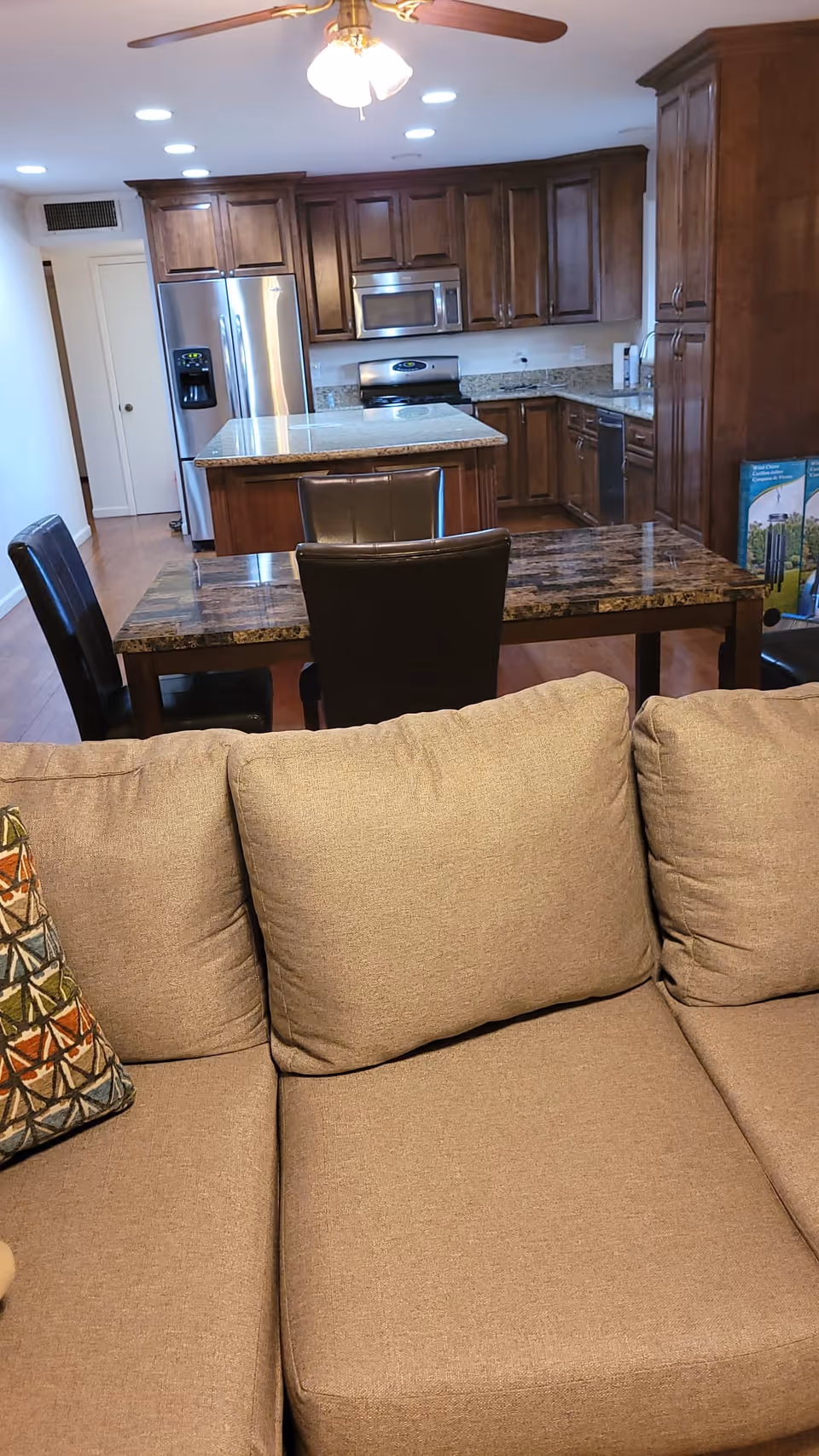 View of a kitchen and dining area from behind a beige couch. The kitchen features wooden cabinets, a stainless steel refrigerator, microwave, stove, and dishwasher. A ceiling fan with lights is visible above, and a dark marble dining table with black chairs is positioned between the couch and kitchen.