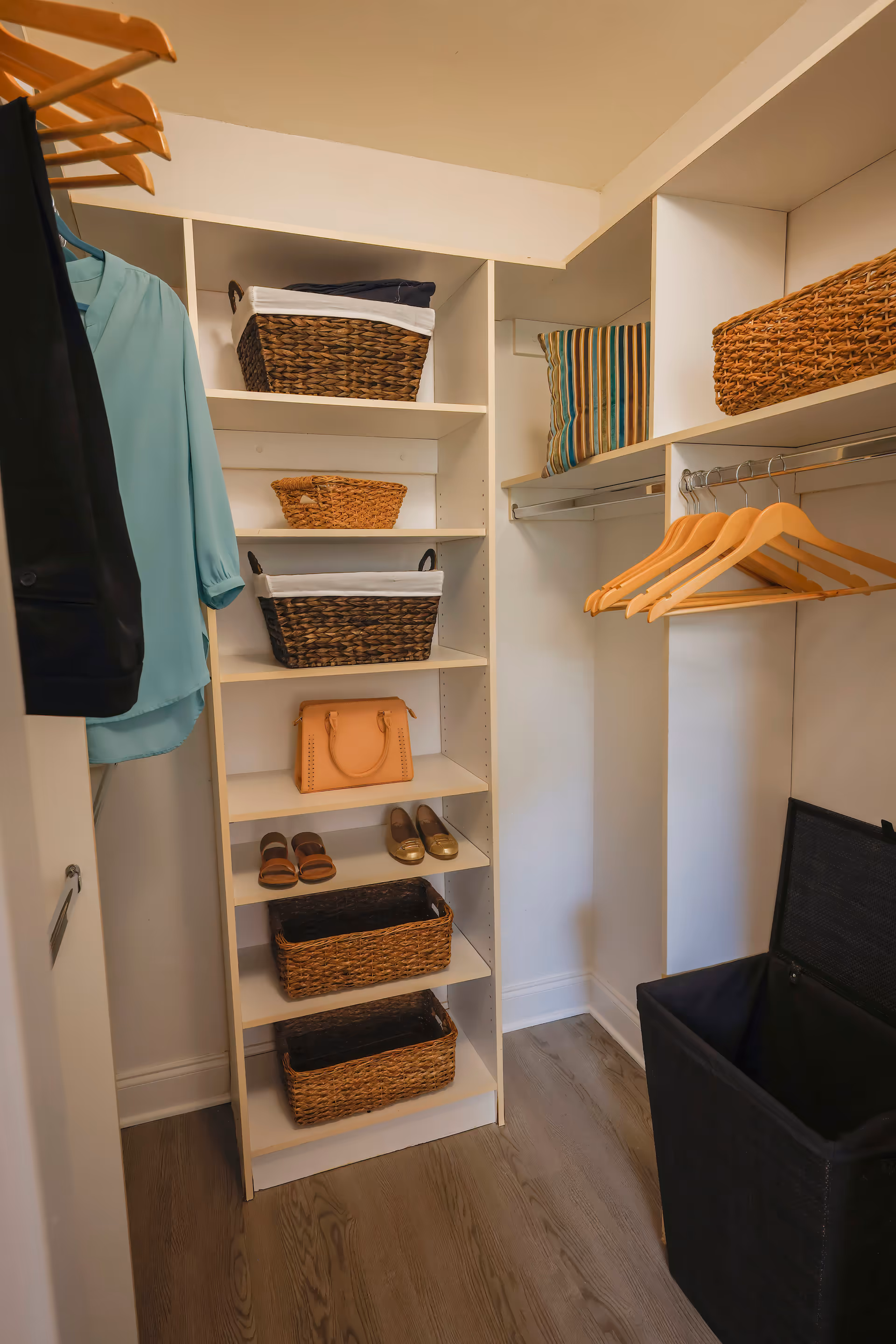 Organized walk-in closet with white shelves holding woven baskets, shoes and a handbag, wooden hangers with hanging clothes, and a black laundry hamper.