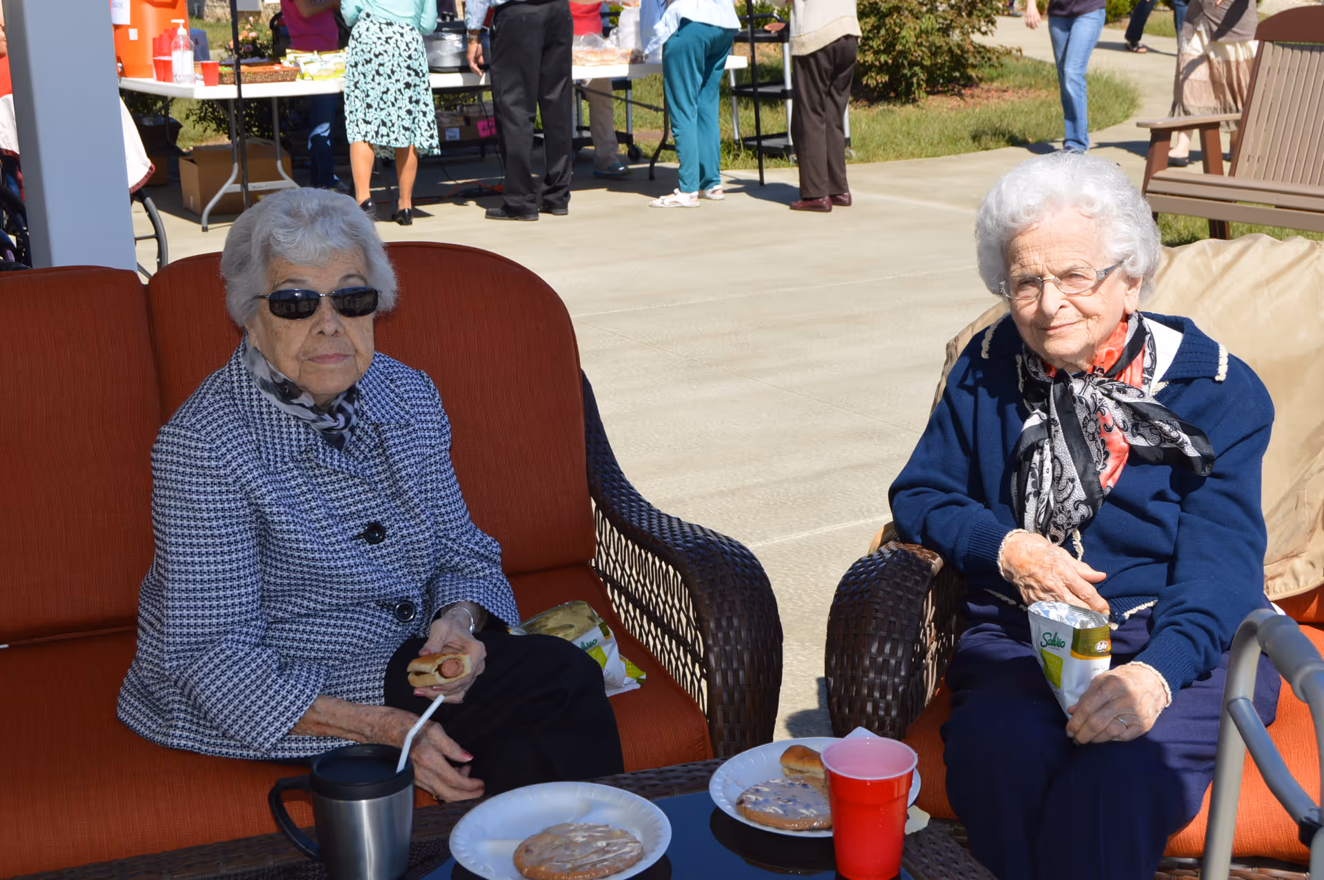 Two elderly women sitting outdoors on cushioned chairs, enjoying snacks and drinks. One woman is wearing sunglasses and a checkered jacket, holding a sandwich, while the other woman is wearing glasses, a navy blue sweater, and a scarf, holding a bag of chips. In the background, other people are standing near a table with refreshments.