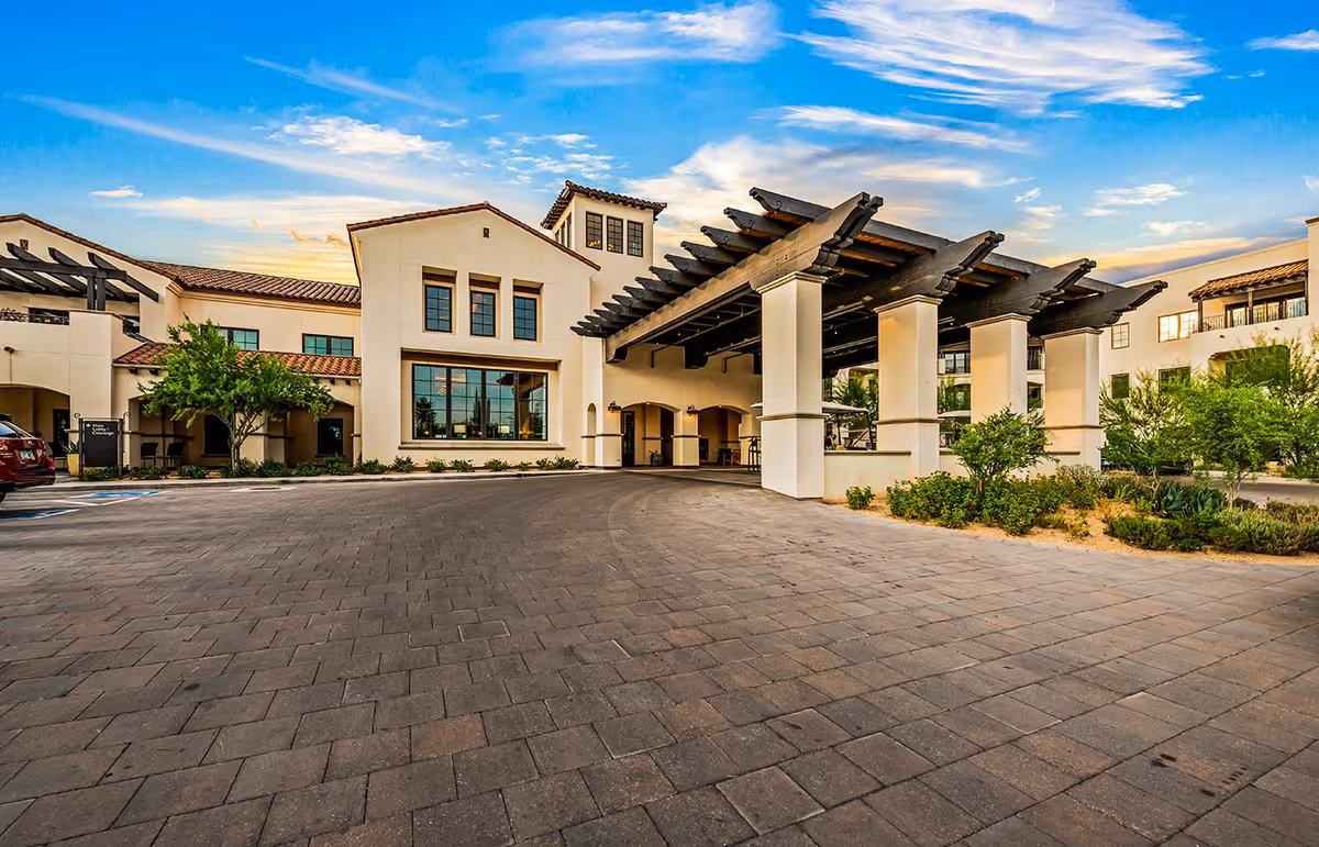 Exterior view of The Hacienda at the Canyon senior living facility showing a large driveway with a covered entrance supported by white pillars and a building with multiple windows and a tiled roof under a partly cloudy blue sky.
