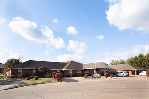 Exterior view of a single-story brick building with a pitched roof under a partly cloudy sky. The building is surrounded by a paved driveway and small landscaped bushes. Several cars are parked near the entrance.