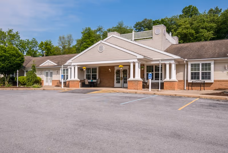 Front entrance of a single-story senior living building with a columned portico, accessible parking spaces, and surrounding trees.