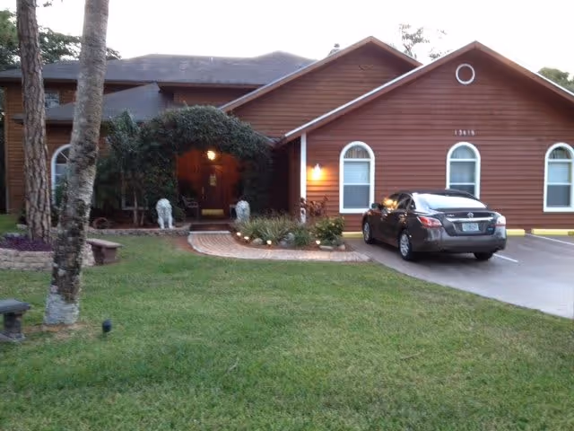 Exterior view of a single-story brown building with white-trimmed windows and a sloped roof. There is a driveway with a parked car on the right side and a green lawn in front. The entrance is partially covered with greenery and lit by a porch light.