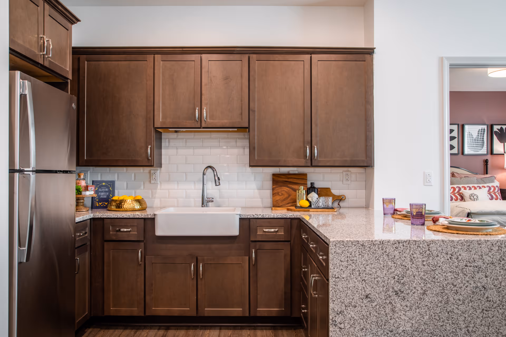 Modern kitchen with dark wooden cabinets, a stainless steel refrigerator, a white farmhouse sink, and a granite countertop. The backsplash is white subway tile. On the countertop, there are decorative items including a basket of lemons, cutting boards, and a few glasses. A dining area with place settings is visible through an open doorway, and part of a bedroom with framed artwork on the wall can be seen beyond that.