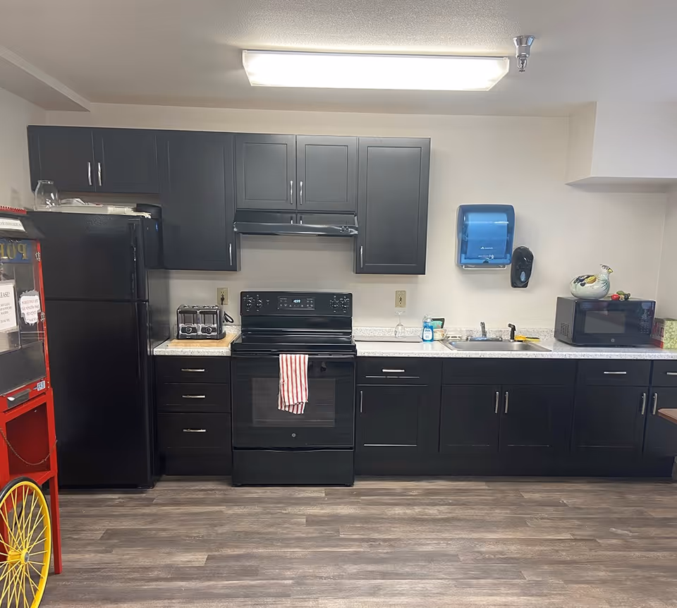 A kitchen area with black cabinets and appliances including a refrigerator, stove with a red and white striped towel hanging on the handle, toaster, microwave, and a sink. There is a blue paper towel dispenser and a black soap dispenser mounted on the wall above the sink. The floor is wood-patterned, and there is a popcorn machine partially visible on the left side.