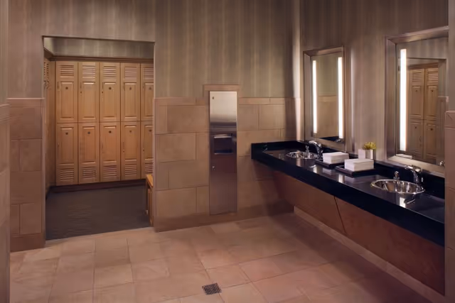 Interior view of a locker room with wooden lockers on the left and a double sink vanity with mirrors and lights on the right. The walls are tiled halfway up and the floor is tiled with large square tiles.