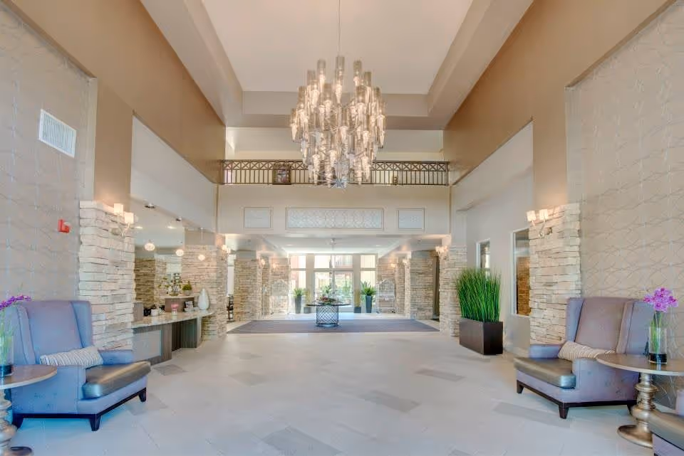 Spacious and elegant lobby area with high ceilings, a large modern chandelier, stone pillar accents, comfortable armchairs with side tables and decorative plants, leading to glass entrance doors.