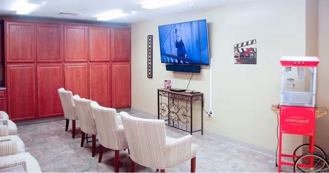 Small assisted-living media room with rows of upholstered chairs facing a wall-mounted TV and a popcorn machine.