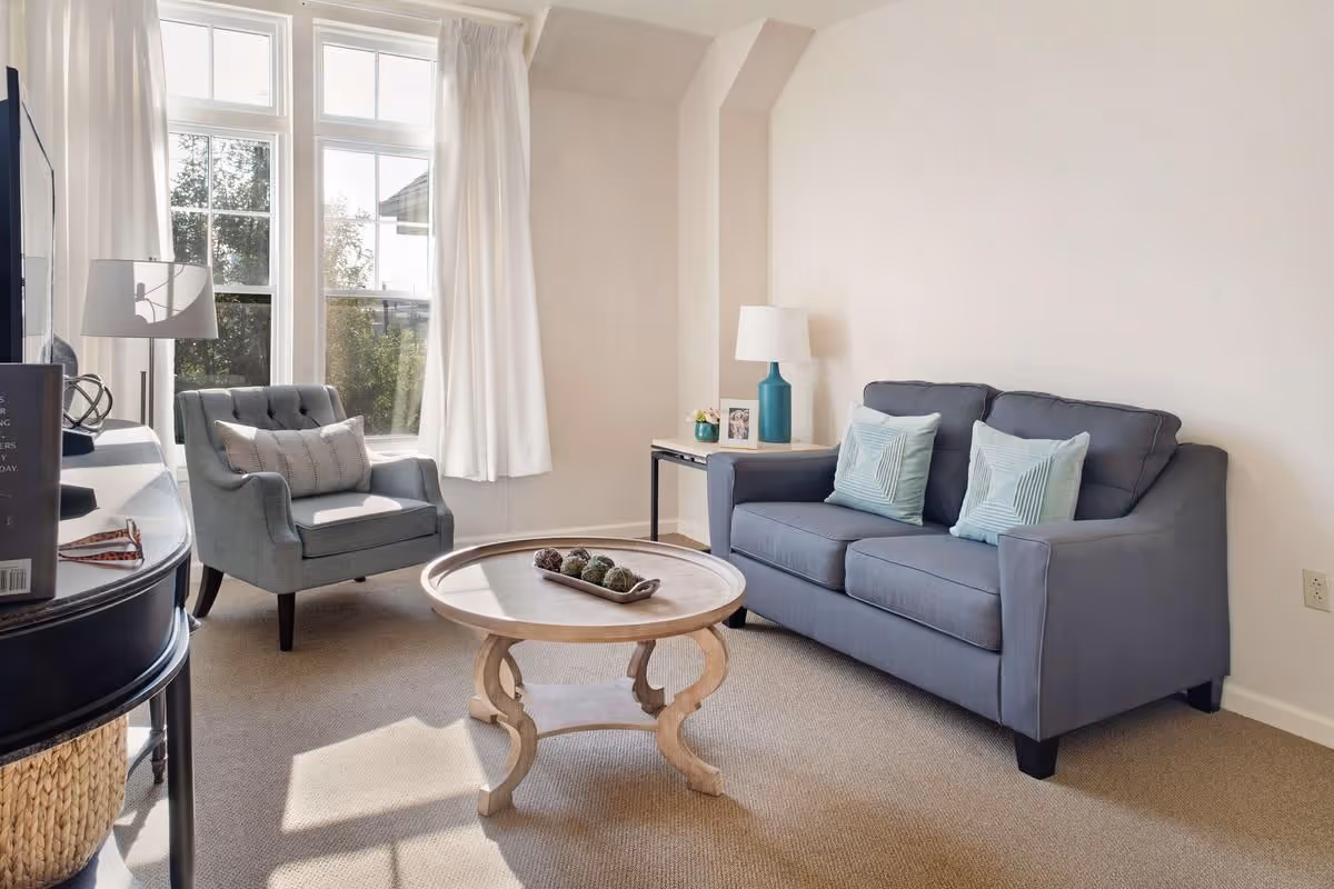 A bright living room with a gray sofa, upholstered armchair, round wooden coffee table, and a large window with white curtains.