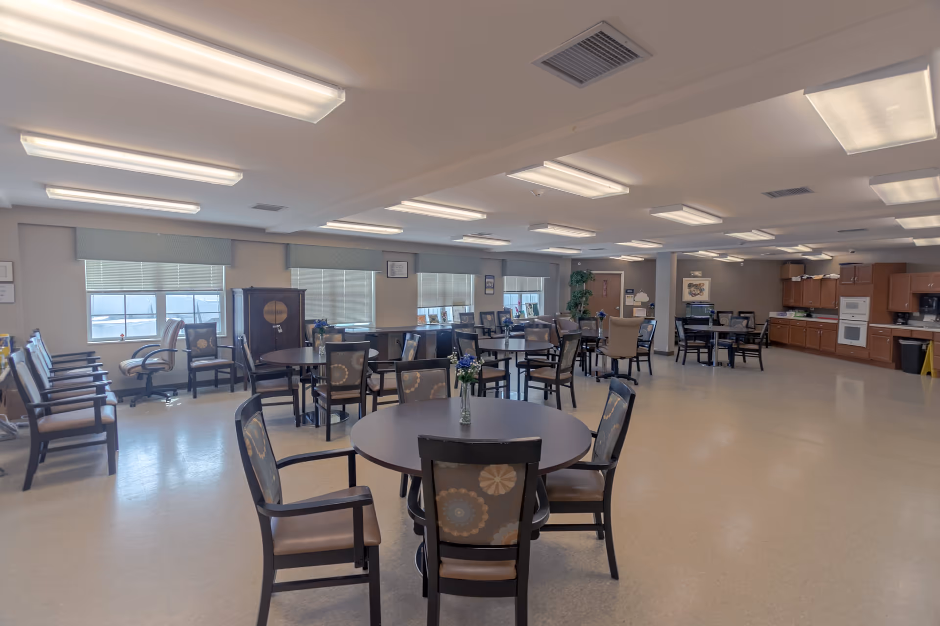 A spacious dining room with multiple round tables and chairs arranged neatly. Each table has a small vase with flowers. The room has large windows with blinds, overhead fluorescent lighting, and a kitchen area with wooden cabinets, a microwave, and an oven in the background.