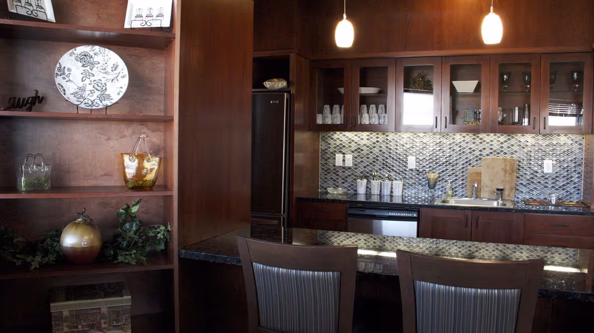 A modern kitchen area with dark wooden cabinets and a mosaic tile backsplash. The kitchen features a sink, dishwasher, and a refrigerator. There are glass-front cabinets displaying cups and bowls. In the foreground, there is a dark granite countertop with two chairs. To the left, there are wooden shelves decorated with a decorative plate, glass containers, a small plant, and a metallic pumpkin.