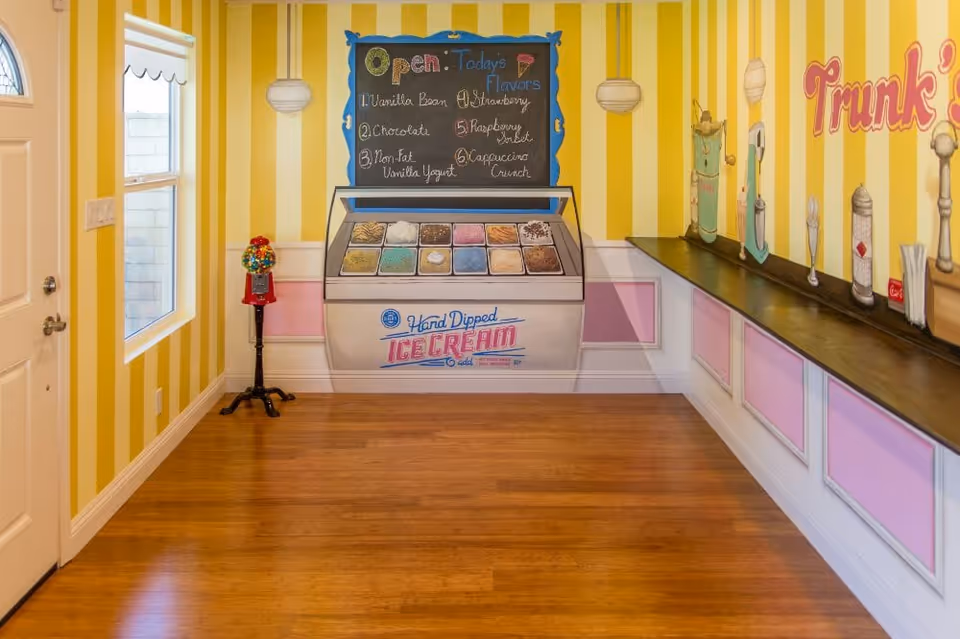 Interior room with yellow and white striped walls featuring a mural of an ice cream display with various flavors listed on a chalkboard. There is a red gumball machine on a stand near a window and a counter along one wall with painted images of condiment dispensers. The floor is wooden and the room is well-lit.