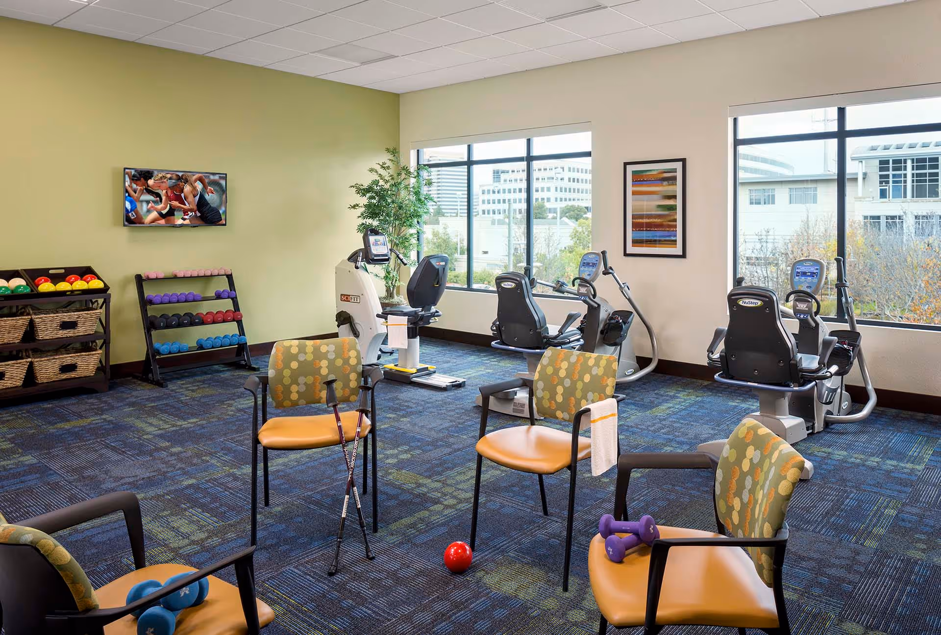 A fitness room with exercise equipment including stationary bikes and a NuStep machine, a rack of colorful dumbbells, and chairs arranged in a circle with small weights and walking sticks on them. Large windows show an urban exterior view, and a TV on the wall displays a running race.