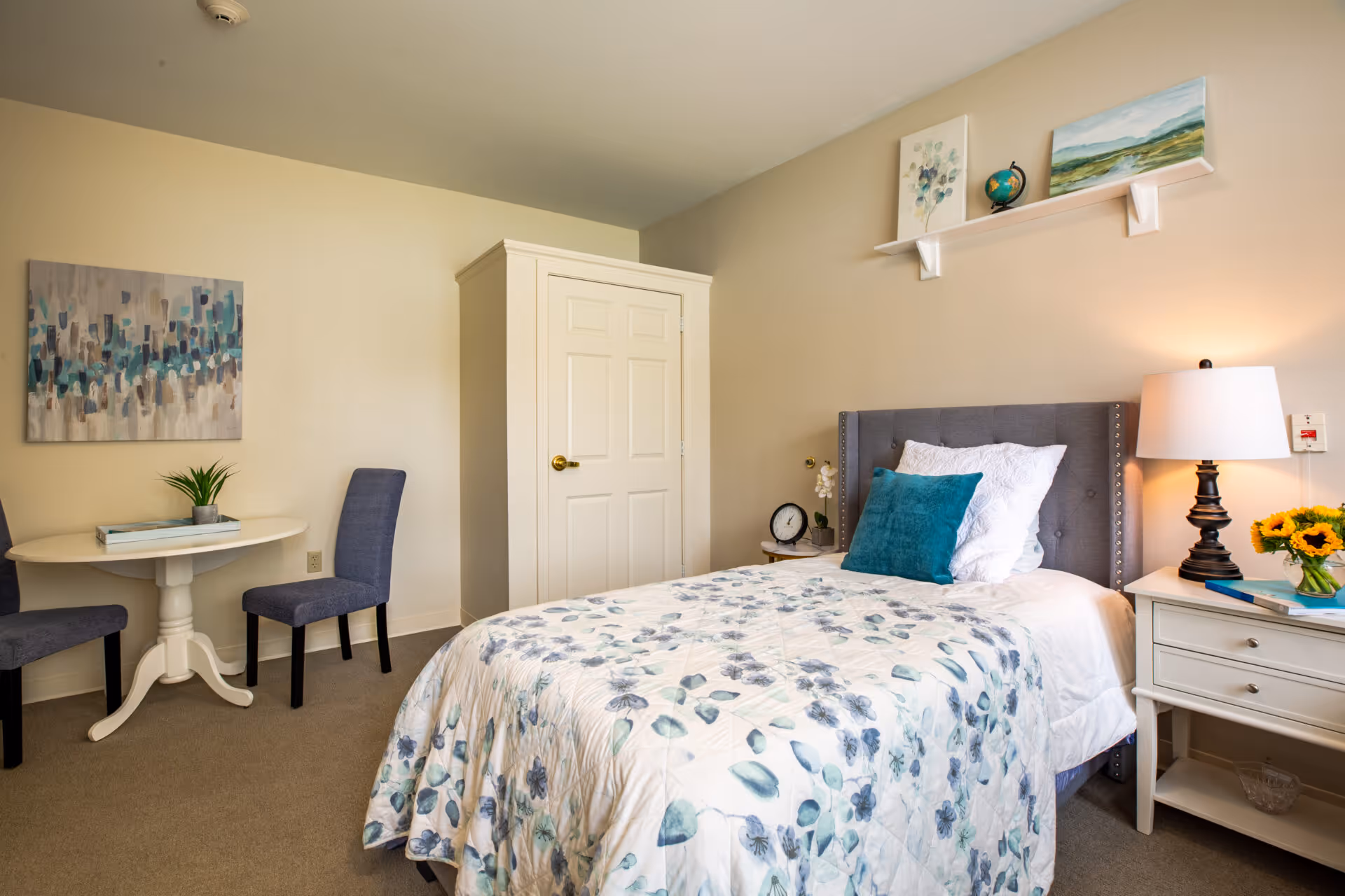 A cozy bedroom with a single bed featuring a floral quilt and teal accent pillow. There is a white nightstand with a lamp, a clock, and a small plant. Above the bed is a white shelf holding decorative items including a globe and framed artwork. To the left, there is a small round white table with two blue upholstered chairs and a piece of abstract art on the wall.