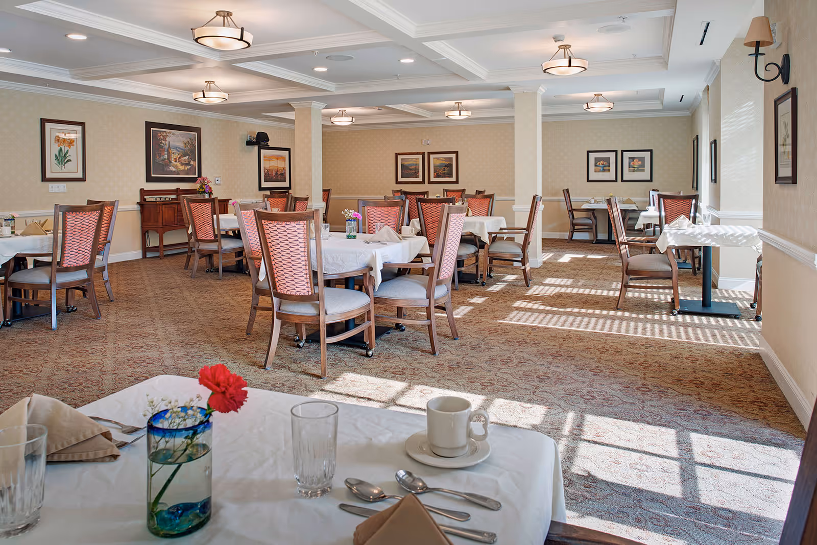A bright dining room with multiple tables covered in white tablecloths and surrounded by wooden chairs with pink cushions. The room has beige walls adorned with framed artwork and a patterned carpet. Sunlight streams through windows, casting shadows on the floor. A table in the foreground has a glass vase with a red flower, a cup and saucer, a glass, and silverware neatly arranged.