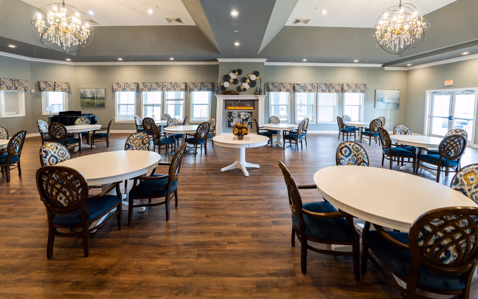 A spacious dining room with multiple round and oval white tables surrounded by wooden chairs with blue and patterned upholstery. The room has large windows with patterned valances, two chandeliers hanging from the ceiling, a fireplace with decorative plates above it, and a floral centerpiece on a central table. The floor is wooden, and there is a piano in the back corner.