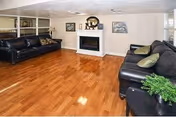 A spacious living room with polished wooden floors, two black leather sofas facing each other, a white fireplace centered on the far wall, and framed artwork above the fireplace. There is a small green plant on a side table next to one of the sofas.