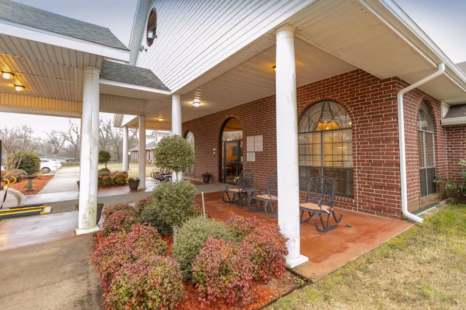 Covered front entrance of a brick senior living facility with white columns, rocking chairs, and landscaped shrubs.