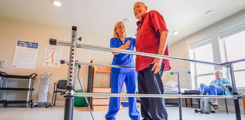 A healthcare professional in blue scrubs assists an elderly man in a red shirt as he practices walking between parallel bars in a rehabilitation or physical therapy room. In the background, an elderly woman sits on an exercise bike near a large window letting in natural light.