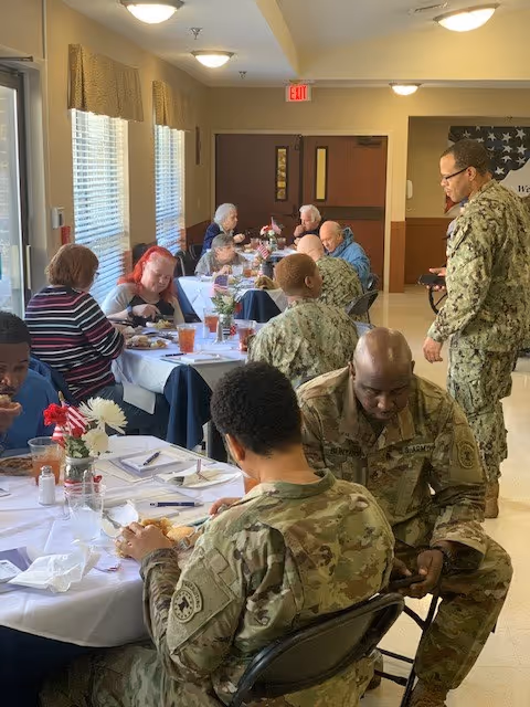A group of people, including several in military uniforms, seated at tables covered with white tablecloths in a dining area. The tables have flowers and drinks, and the people appear to be eating and conversing. The room has windows with curtains and a door at the back.
