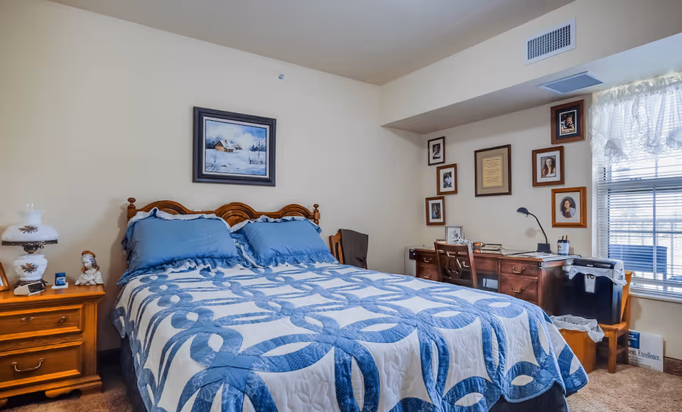 A cozy bedroom with a wooden bed frame and a blue and white patterned quilt. Two blue pillows rest on the bed. To the left is a wooden nightstand with a decorative lamp and small figurines. On the right side of the room is a wooden desk with a chair, books, and framed photos on the wall above it. A window with white curtains allows natural light into the room.