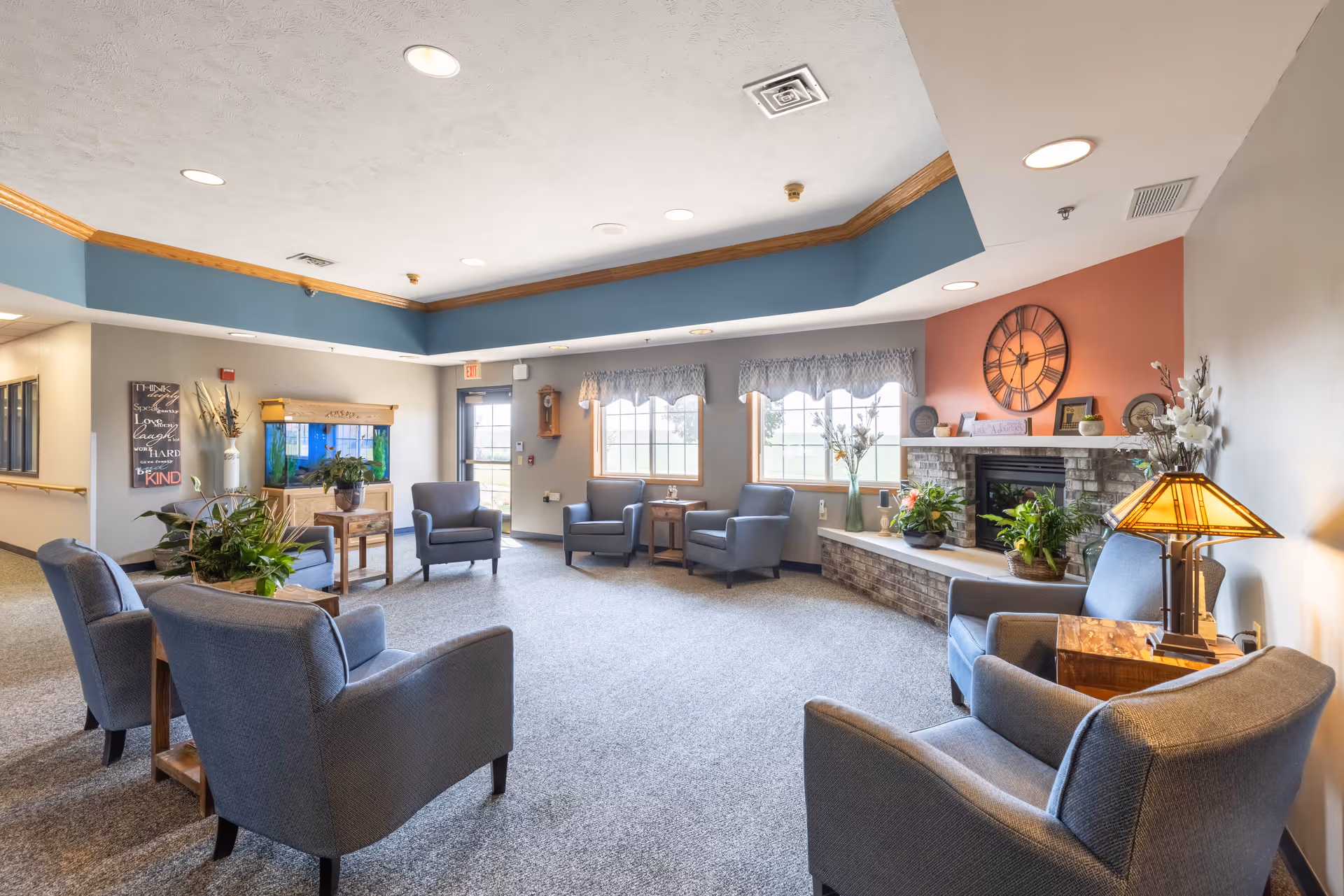 A spacious senior living facility common area with multiple gray upholstered armchairs arranged in a circle on a carpeted floor. The room features a brick fireplace with plants and decorative items on the mantel, large windows with valances letting in natural light, a large wall clock above the fireplace, and a fish tank on a wooden stand. The ceiling has recessed lighting and a blue accent border with wood trim.