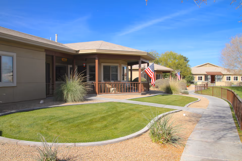Front exterior of a single-story senior living community with a covered porch, American flags, curved walkway and landscaped lawn.