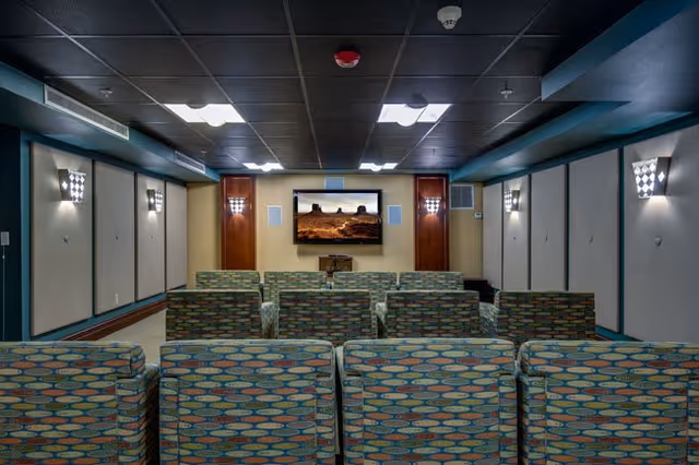 A small theater room with multiple rows of patterned cushioned chairs facing a large flat-screen TV mounted on the wall. The room has a dark ceiling with recessed lighting and wall sconces on the side walls.