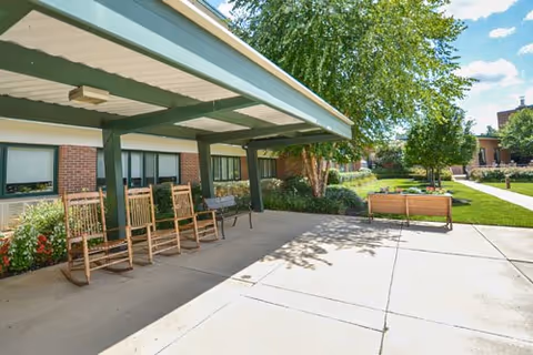 Outdoor patio area at a senior living facility with rocking chairs and benches under a covered walkway, surrounded by greenery and trees on a sunny day.