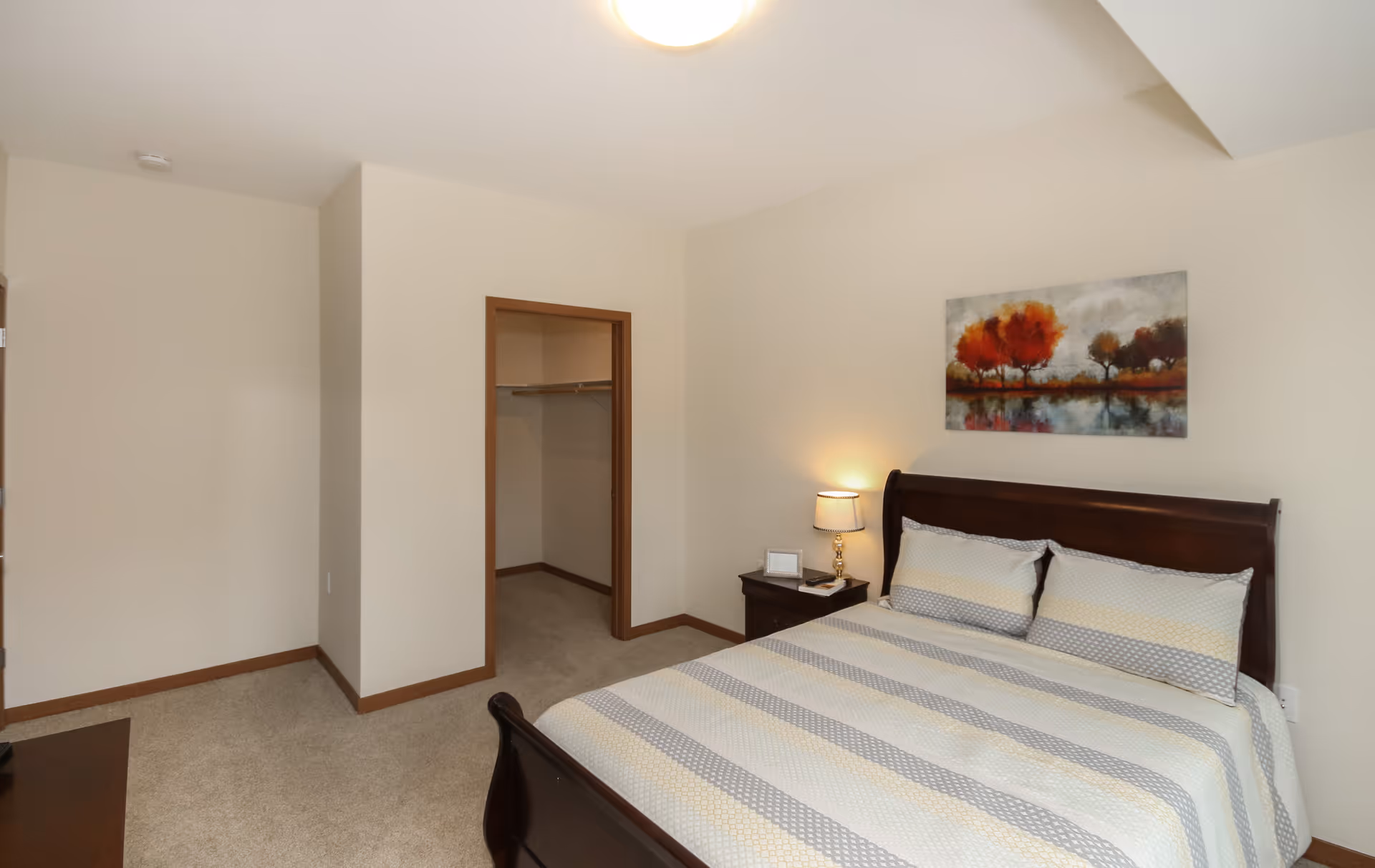 A bedroom with a neatly made bed featuring a striped bedspread in shades of gray, white, and yellow. There is a wooden nightstand with a lamp, a picture frame, and a book next to the bed. Above the bed hangs a painting of trees with red and orange foliage reflected in water. The room has beige walls, carpeted floor, and an open walk-in closet with a wooden frame.