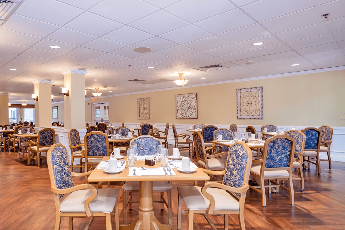 A spacious dining room with multiple wooden tables and chairs featuring blue patterned upholstery. Each table is set with white cups, glasses, silverware, and placemats. The room has warm yellow walls adorned with decorative framed art and is well-lit with ceiling lights and wall sconces. The floor is wooden, and the ceiling has a grid pattern with recessed lighting.