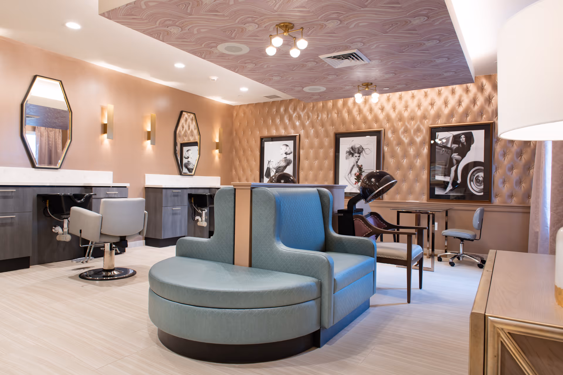 A stylish salon area in a senior living facility featuring two blue salon chairs with hair dryers, two gray styling chairs in front of mirrors mounted on a peach-colored wall, and framed black-and-white photographs on a tufted wall. The ceiling has a decorative pattern and modern light fixtures, with a light-colored tiled floor.